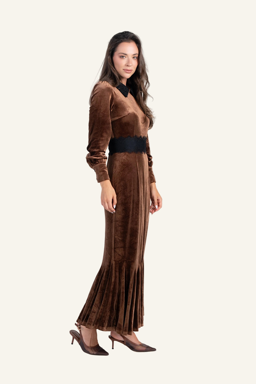 A woman modeling a long-sleeved brown velvet maxi dress with a black collar, black waistband, and black pointed-toe heels.