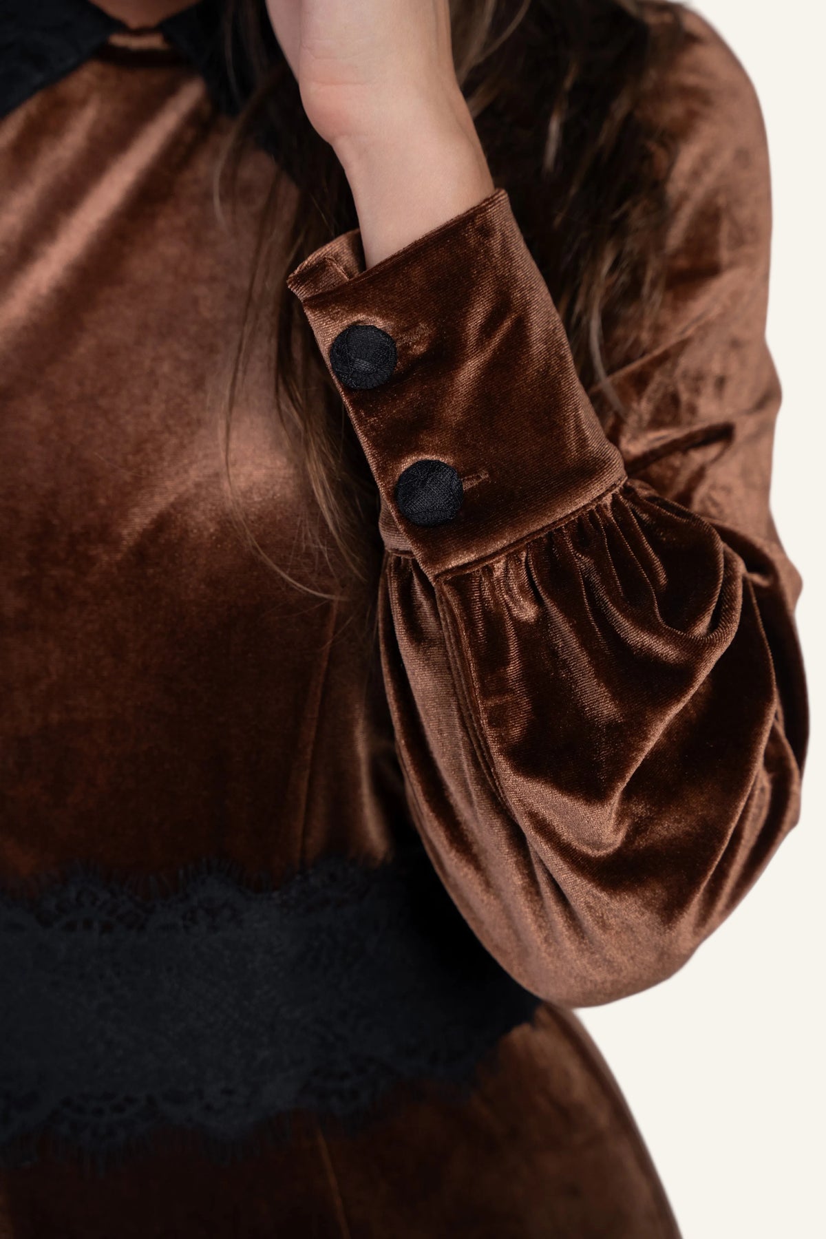 Close-up of a brown velvet long-sleeve top or dress with black buttons on the cuff, gathered sleeves, and a black lace band near the waist, shown on a person.