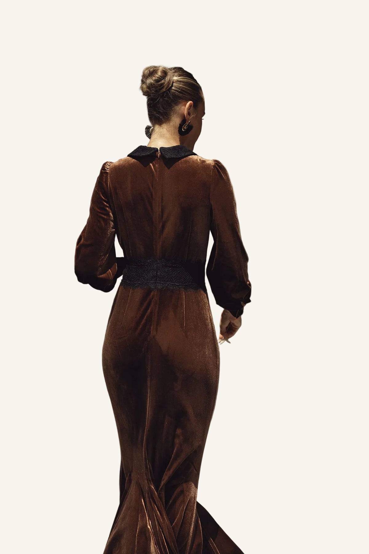 A woman viewed from behind wearing a long brown velvet gown with a black collar and a wide black waistband.