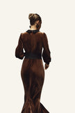 A woman viewed from behind wearing a long brown velvet gown with a black collar and a wide black waistband.