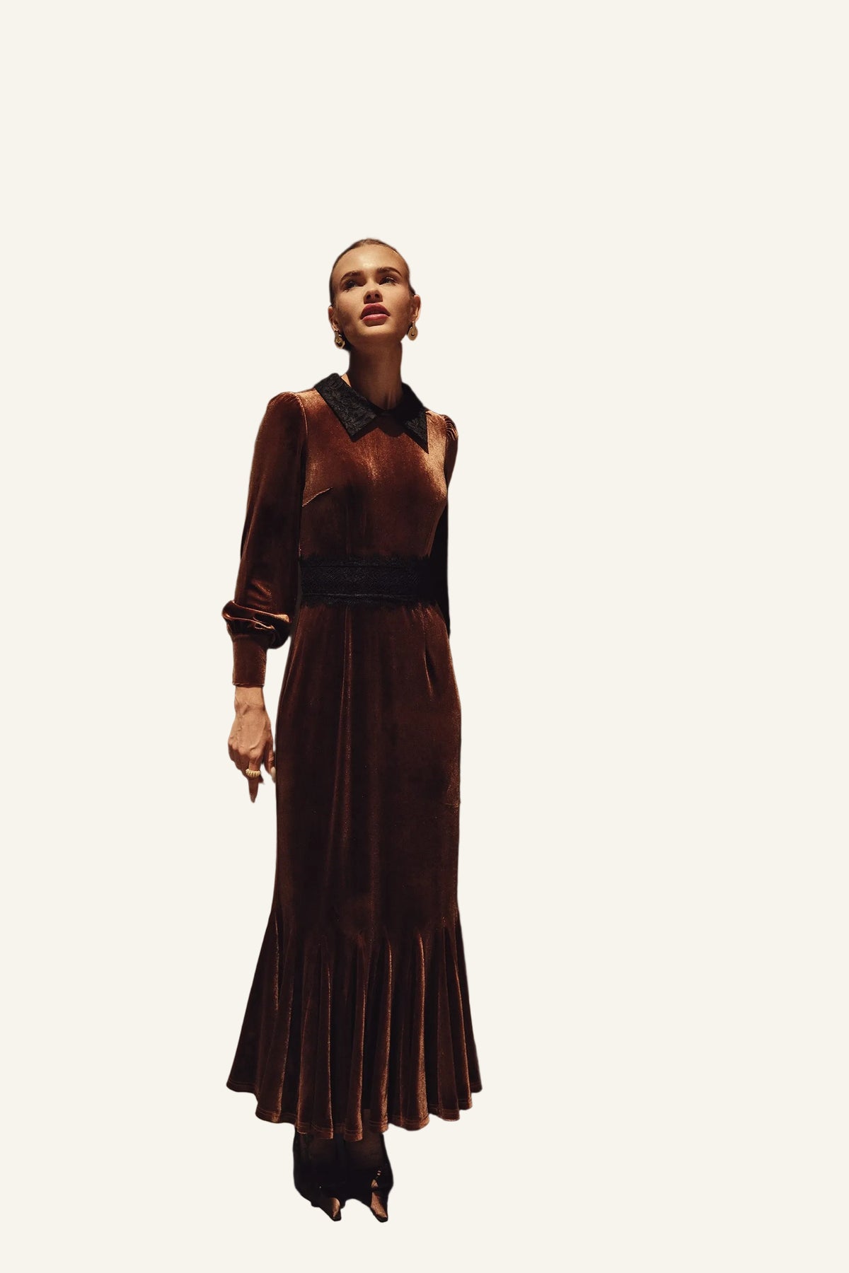 A woman wearing a long brown velvet dress with a black collar and a wide black waistband. The dress has long sleeves and a flared hem, photographed in an urban outdoor setting with buildings and greenery in the background.