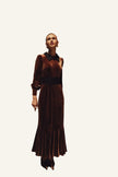 A woman wearing a long brown velvet dress with a black collar and a wide black waistband. The dress has long sleeves and a flared hem, photographed in an urban outdoor setting with buildings and greenery in the background.