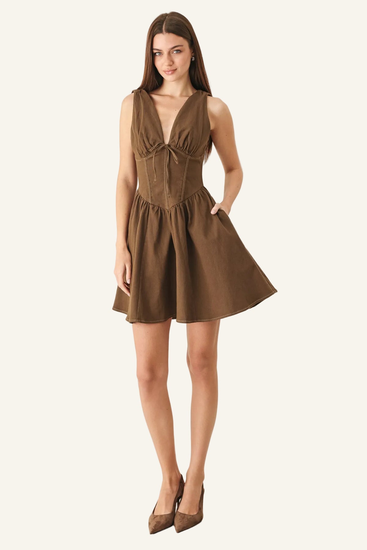 A woman modeling a sleeveless brown mini dress with a deep V-neckline, gathered bust, and flared skirt, paired with brown pointed-toe heels against a plain light gray studio background.