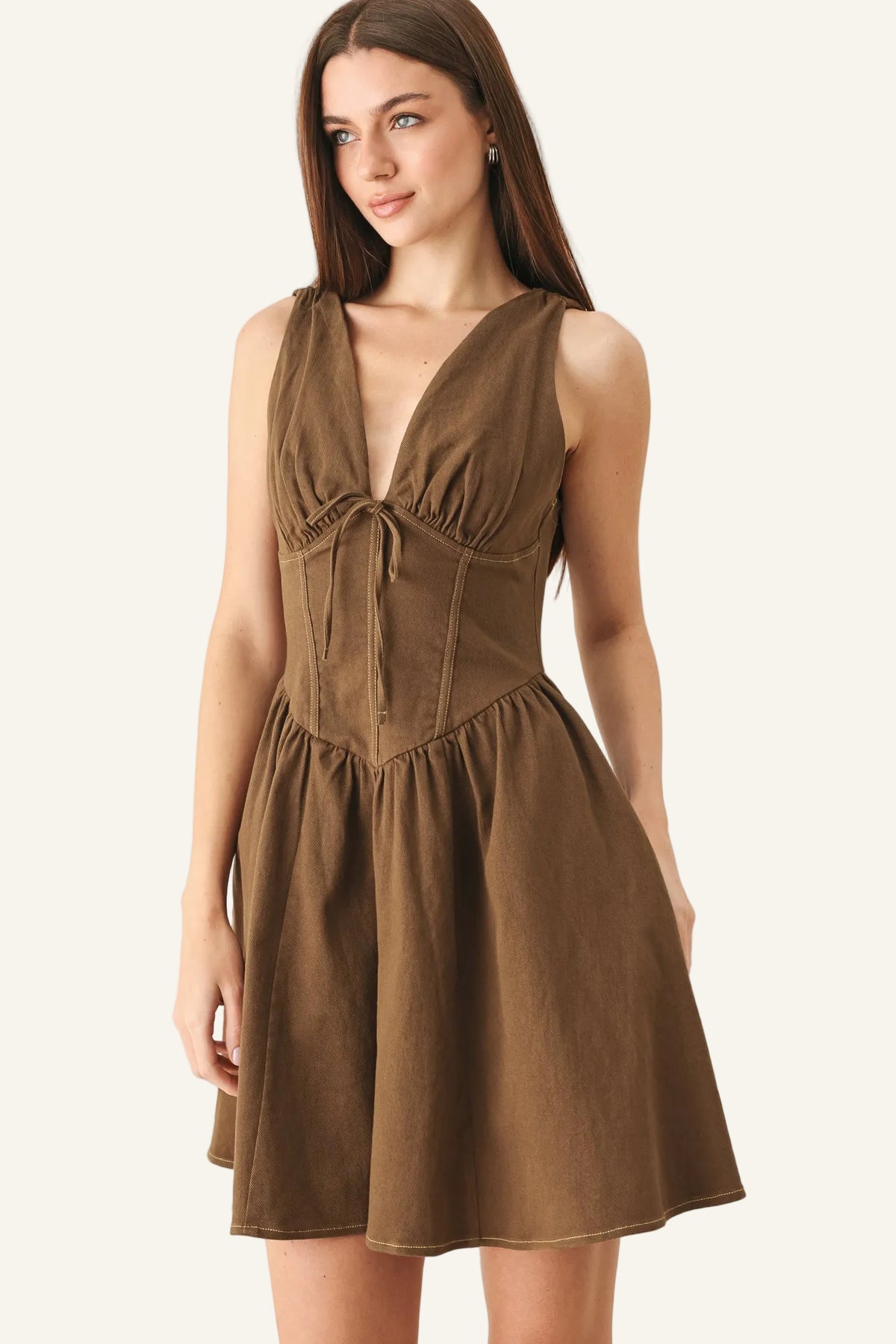 A woman modeling a sleeveless brown mini dress with a deep V-neckline, gathered bust, and flared skirt against a plain light gray studio background.