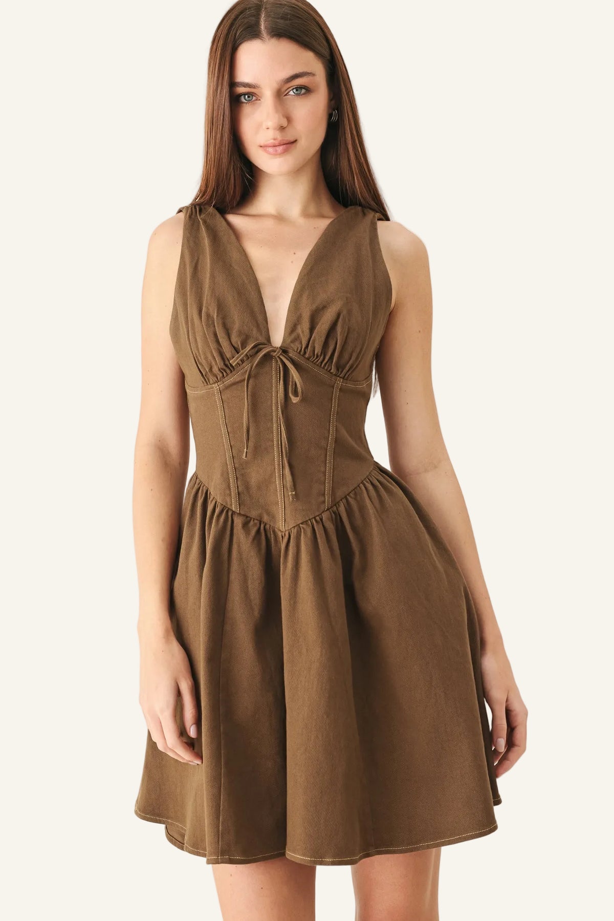 A woman modeling a sleeveless brown mini dress with a deep V-neckline, gathered bust, and flared skirt against a plain light gray studio background.