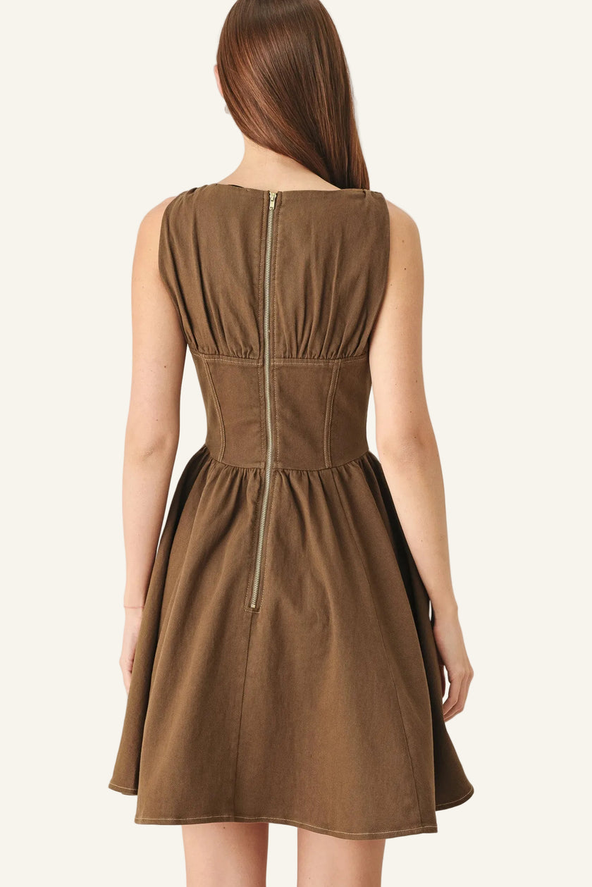 A woman shown from the back wearing a sleeveless brown dress with a gathered waist and a central zipper, photographed against a plain light gray studio background.