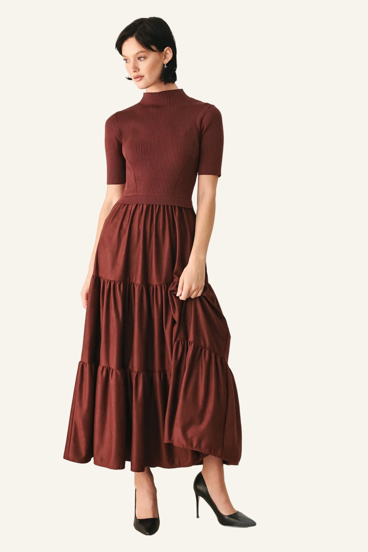A woman modeling a short-sleeve mock-neck midi dress in a deep burgundy color, paired with black high heels.
