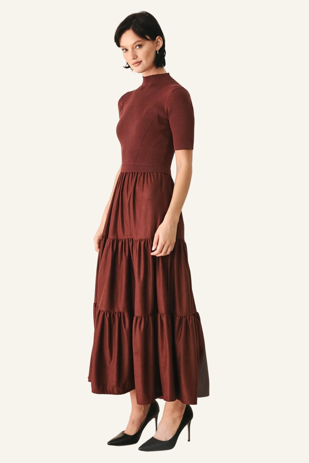 A woman modeling a rust-brown tiered midi dress with short sleeves and a mock neck, paired with black pointed-toe heels.