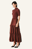 A woman modeling a rust-brown tiered midi dress with short sleeves and a mock neck, paired with black pointed-toe heels.