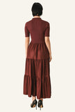 A woman viewed from the back wearing a short-sleeve, high-neck, tiered maxi dress in a deep burgundy or maroon shade, paired with black high heels.