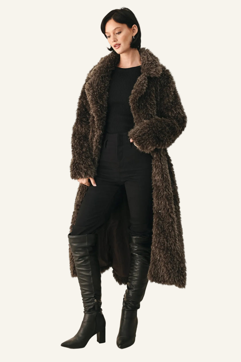 A model wearing a long, dark brown shaggy faux fur coat over a black outfit with black leather knee-high boots, photographed against a plain light gray studio background.