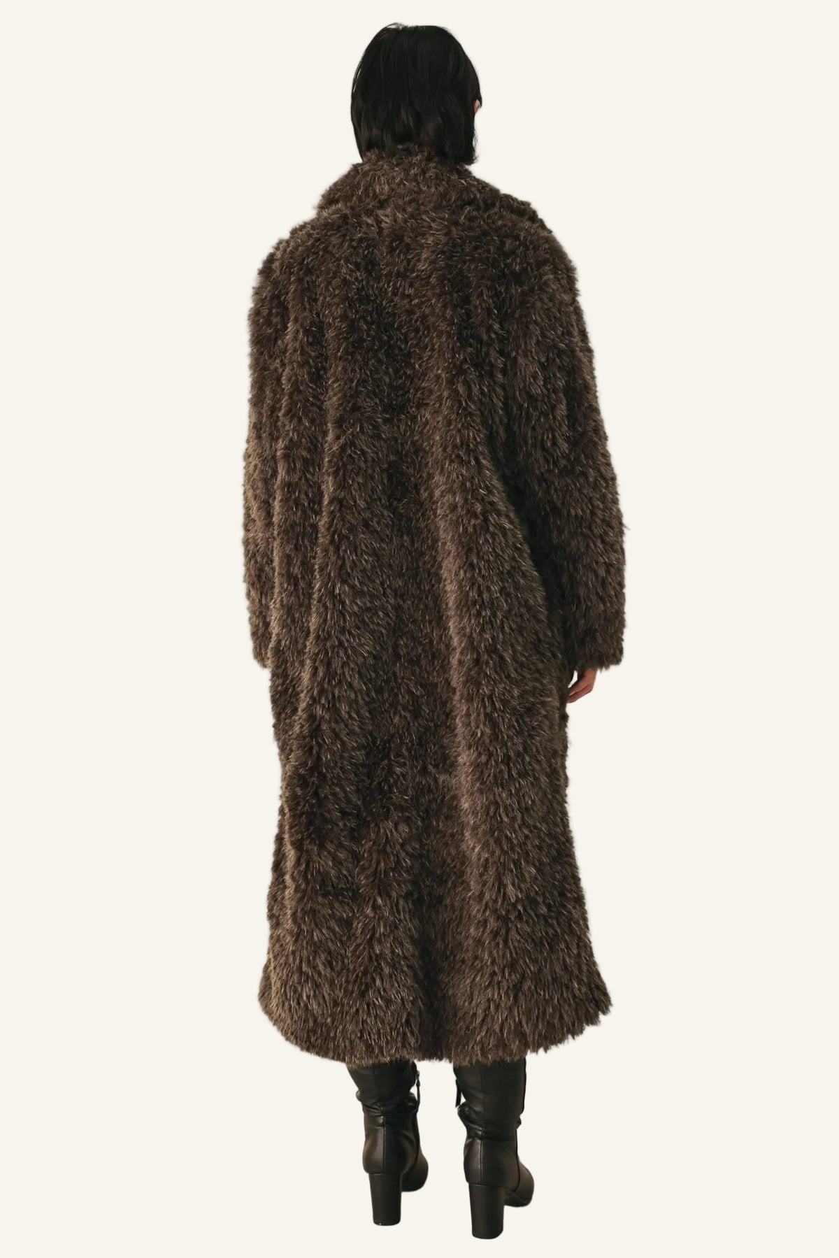 A person viewed from the back wearing a long, shaggy brown faux-fur coat over black knee-high boots, standing against a plain light background.