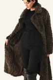 A woman wearing a long, dark brown shaggy faux-fur coat over a black ribbed top, black pants, and black boots, standing against a plain white background.