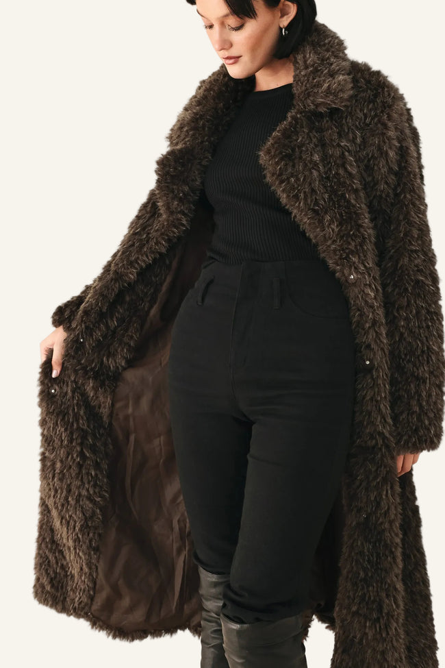 A woman wearing a long, dark brown shaggy faux-fur coat over a black ribbed top, black pants, and black boots, standing against a plain white background.