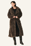 A woman modeling a long, shaggy brown faux fur coat over a black outfit and black knee-high boots, photographed against a plain light studio background.