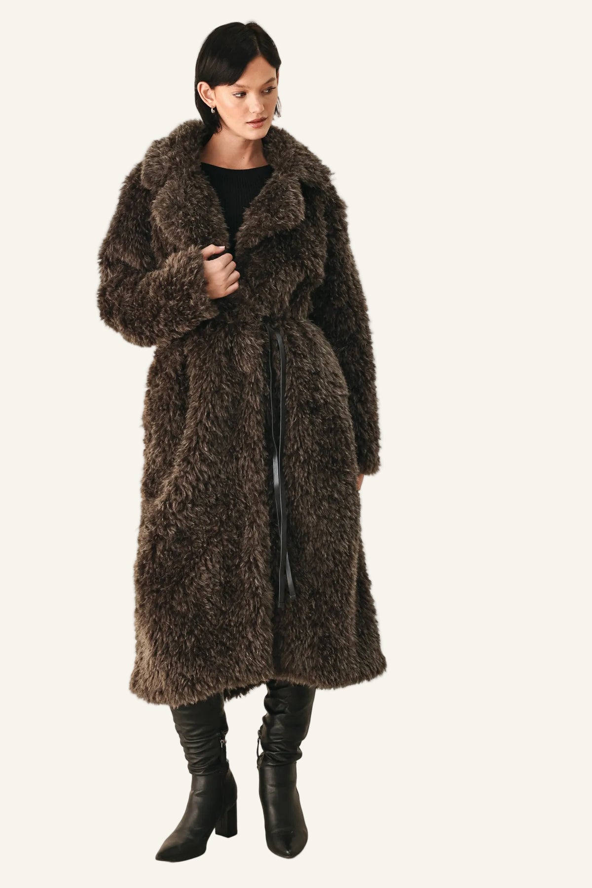 A model wearing a long, dark brown shaggy faux fur coat over a black top and black leather boots, standing against a plain off-white studio background.