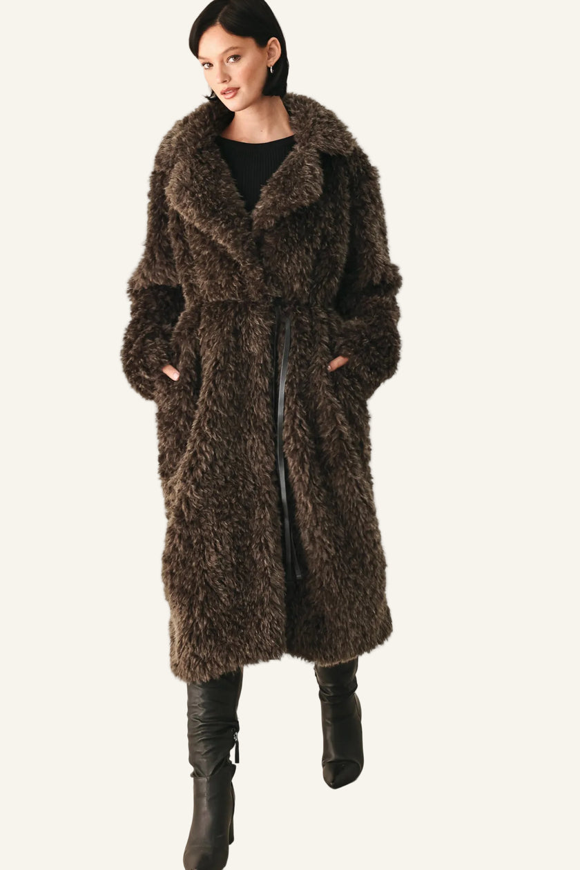 A woman modeling a long, shaggy brown faux fur coat with a wide collar, worn over a black top and black knee-high boots, against a plain white studio background.