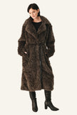 A woman modeling a long, shaggy brown faux fur coat with a black top and black heeled boots, standing against a plain light gray studio background.