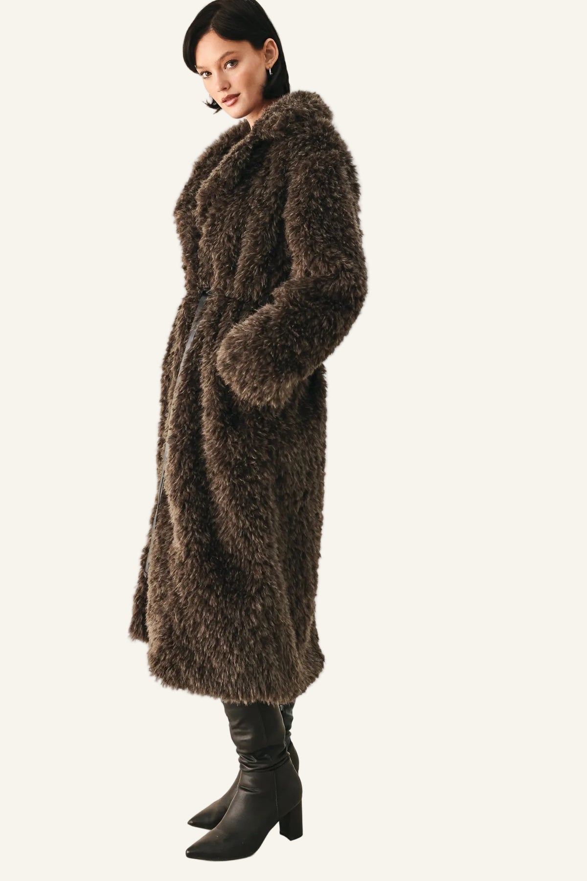 A woman modeling a long, shaggy brown faux fur coat with black leather ankle boots, standing against a plain off-white studio background.