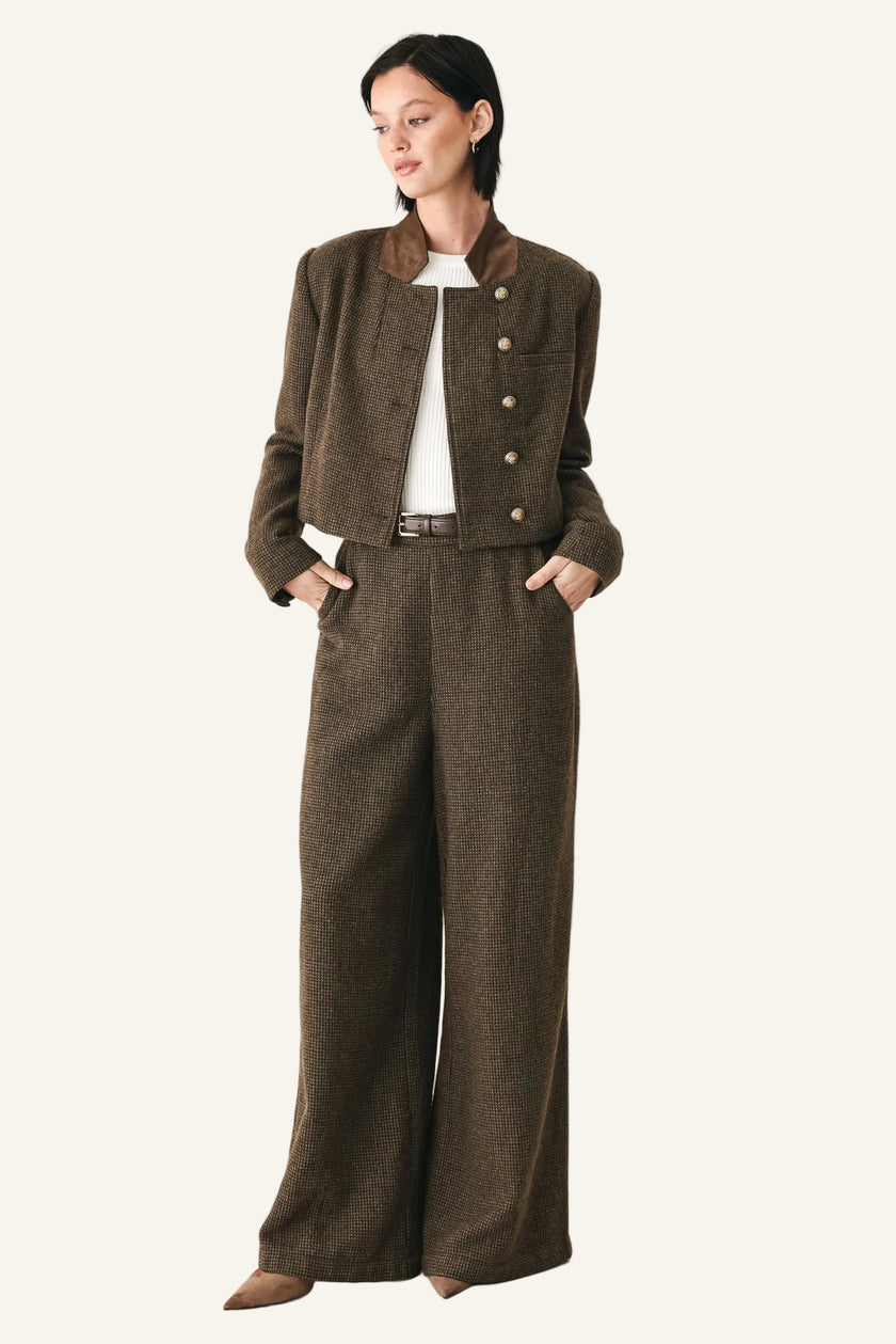 A model stands against a plain white studio background wearing a matching brown double-breasted jacket and wide-leg trousers set with a white top underneath.