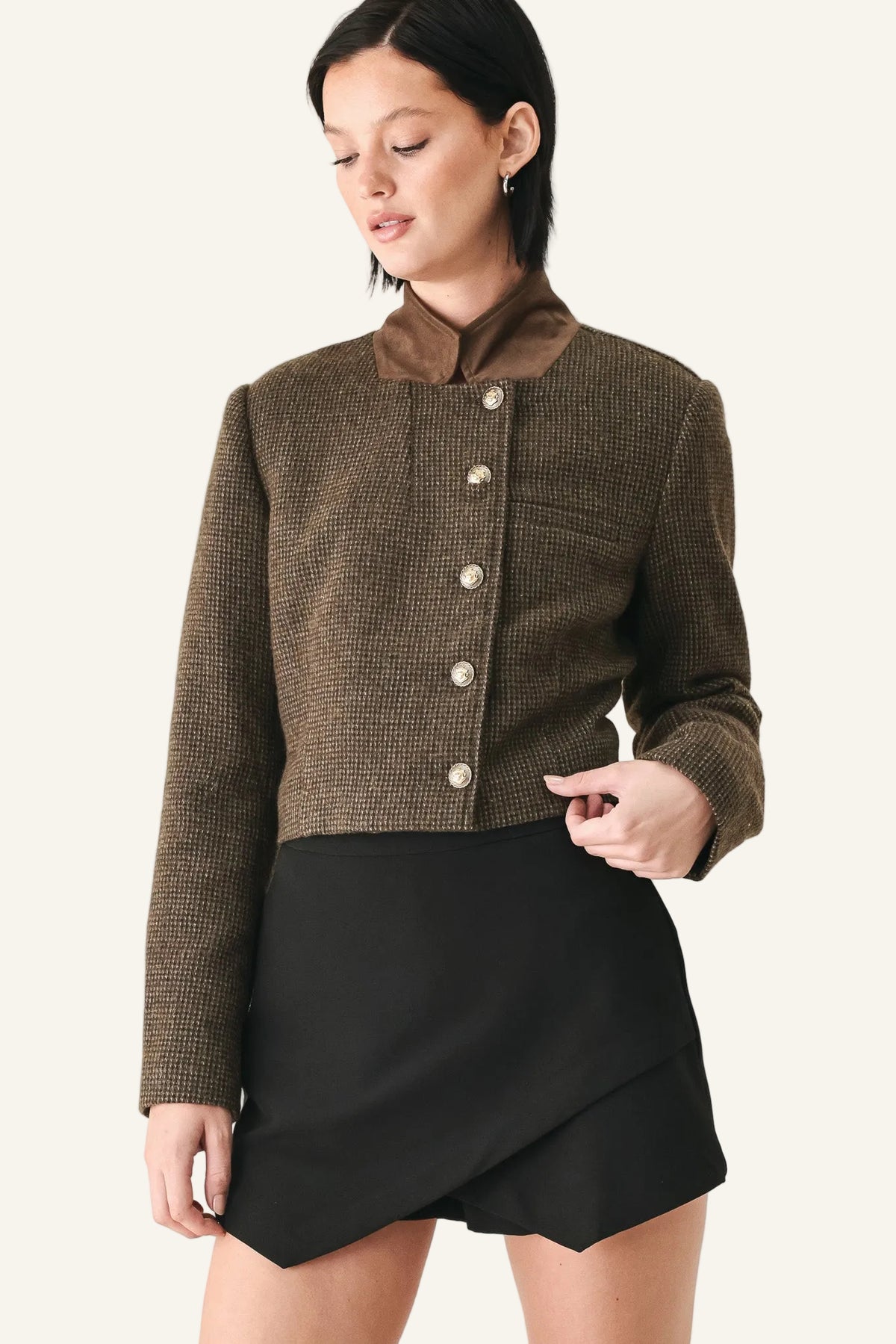 A woman wearing a cropped brown tweed jacket with a high brown collar and silver buttons, paired with a black wrap-style mini skirt.