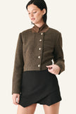 A woman wearing a cropped brown tweed jacket with a high brown collar and silver buttons, paired with a black wrap-style mini skirt.