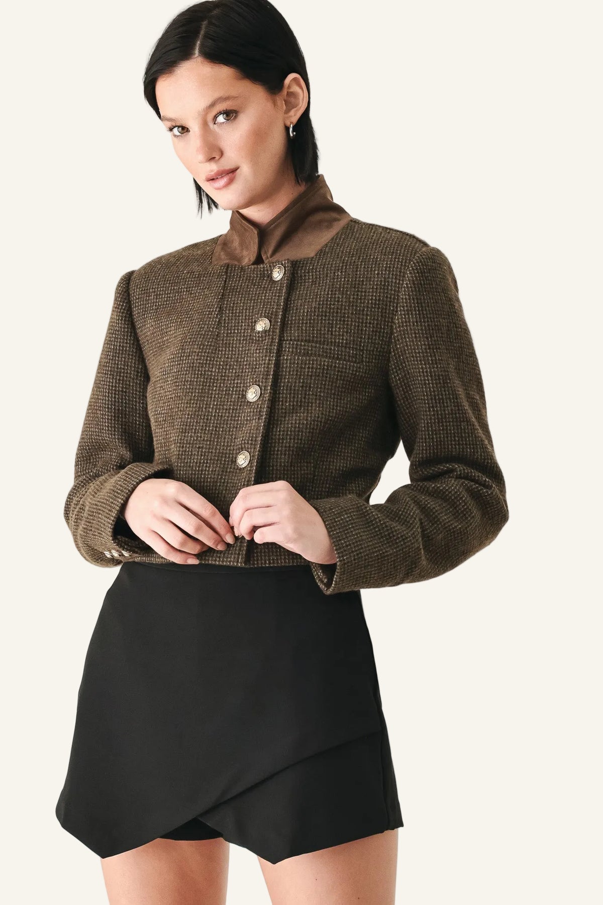 A model wearing a cropped brown tweed jacket with a high collar and button front, paired with a black wrap-style mini skirt.