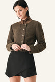 A model wearing a cropped brown tweed jacket with a high collar and button front, paired with a black wrap-style mini skirt.