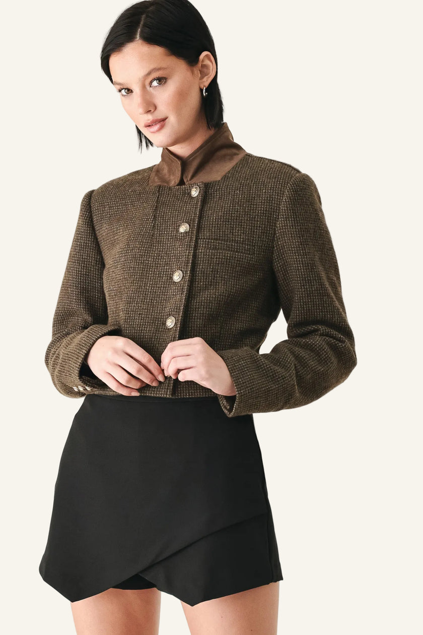A model wearing a cropped brown tweed jacket with a high collar and button front, paired with a black wrap-style mini skirt.