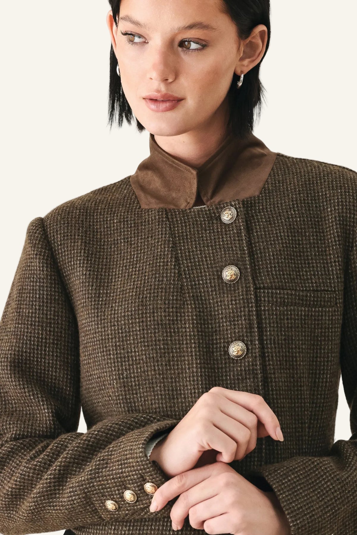 A close-up of a model wearing a brown textured tweed jacket with a contrasting solid brown collar and decorative gold-tone buttons.