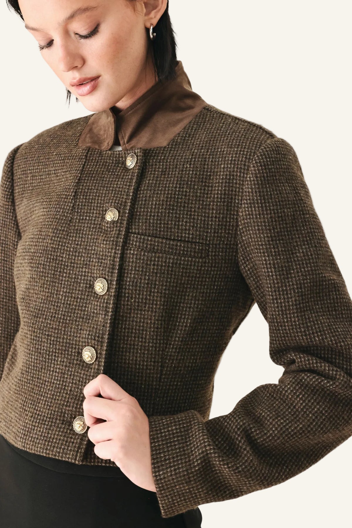 A close-up of a model wearing a cropped brown tweed jacket with a high brown collar and gold-tone buttons.