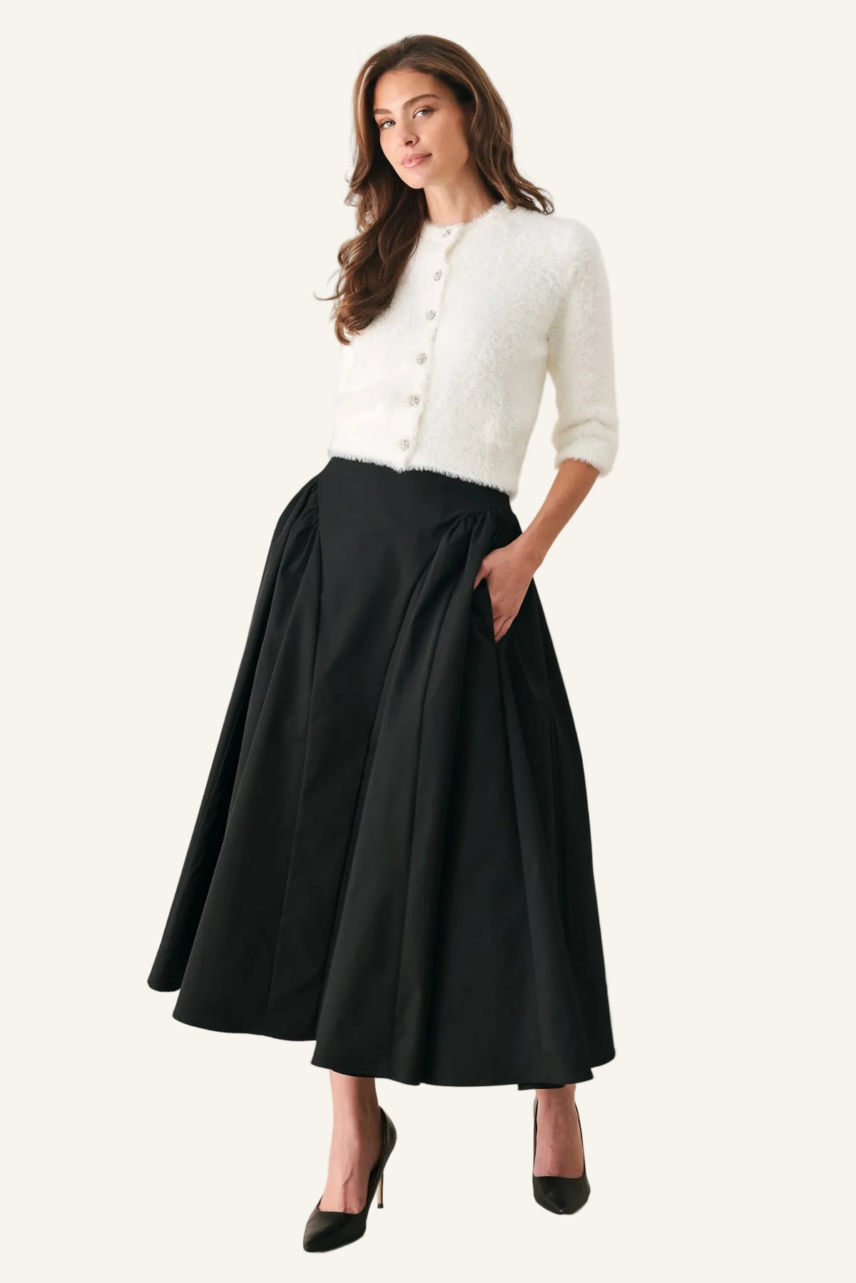 A woman modeling a white textured knit cardigan and a black pleated midi skirt with pockets, paired with black pointed-toe heels, against a plain light gray studio background.