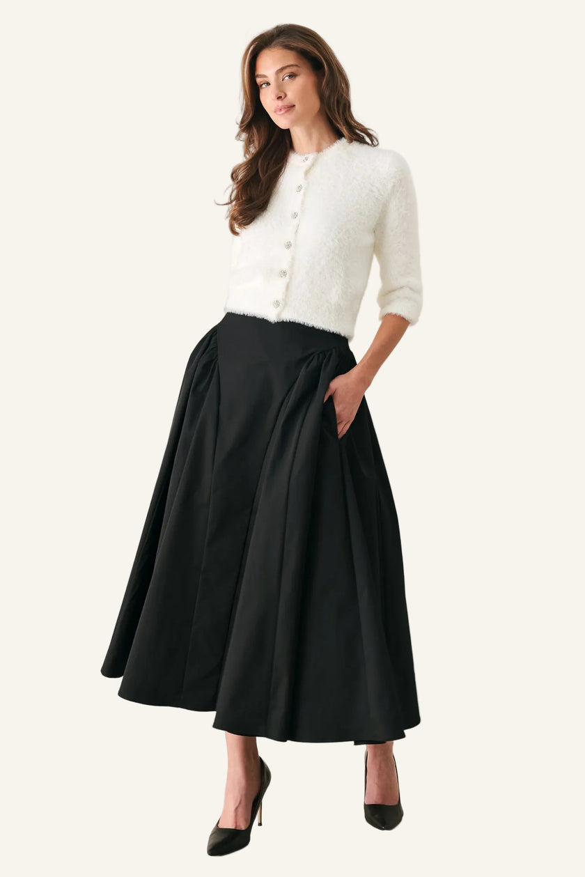 A woman modeling a white textured knit cardigan and a black pleated midi skirt with pockets, paired with black pointed-toe heels, against a plain light gray studio background.