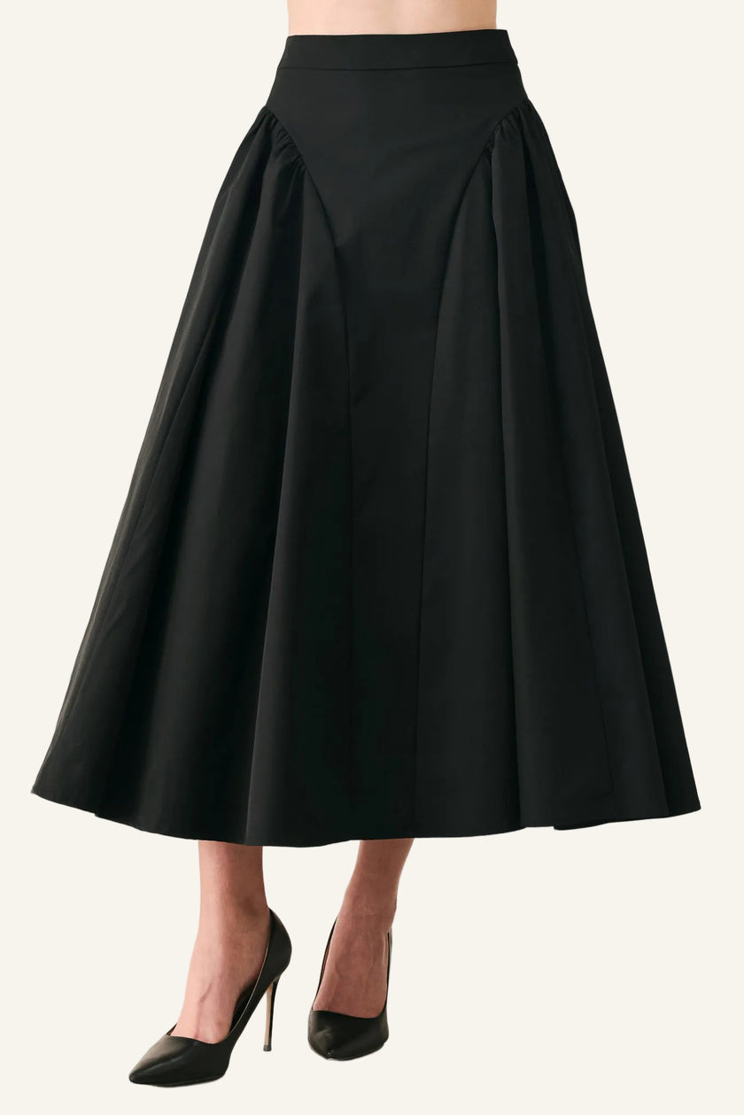 A black high-waisted A-line midi skirt with pleated detailing, shown on a model wearing black pointed-toe stiletto heels against a light gray studio background.
