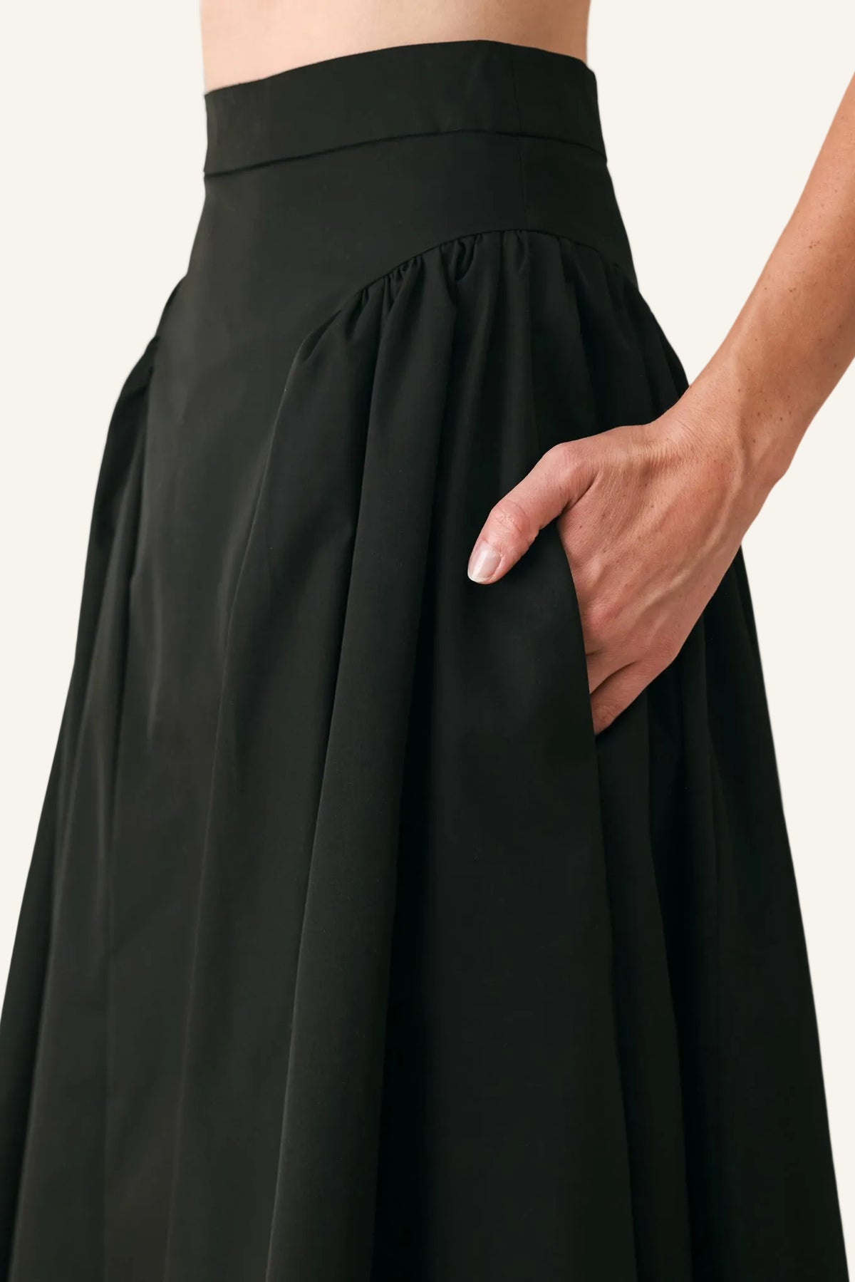 Close-up of a black high-waisted pleated skirt with a side pocket, shown on a model against a light gray background.