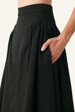 Close-up of a black high-waisted pleated skirt with a side pocket, shown on a model against a light gray background.