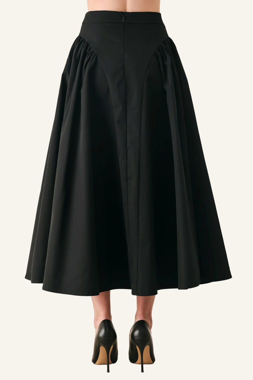 A black high-waisted A-line midi skirt with dramatic gathered side pleats, shown from the back on a model wearing black pointed-toe heels against a light gray studio background.