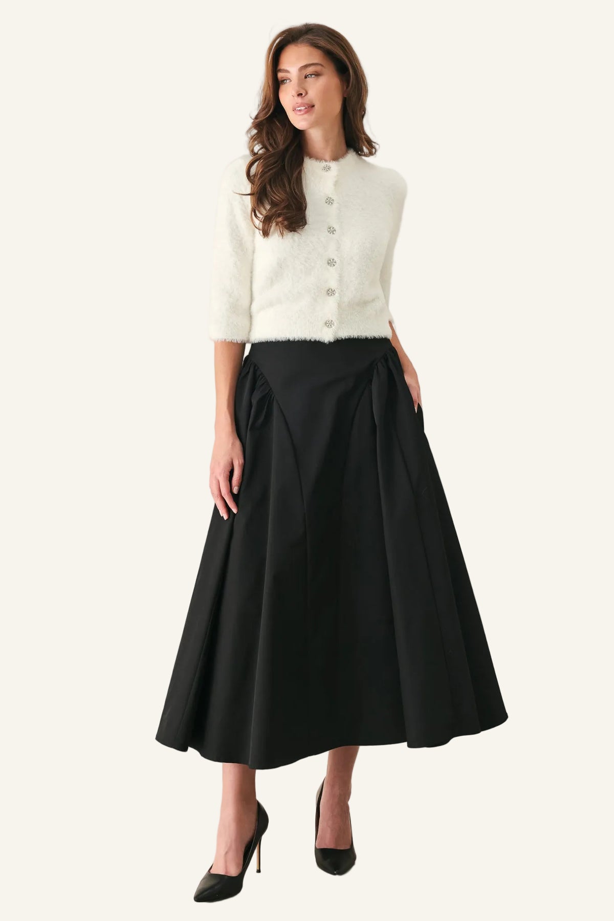 A woman modeling a cream textured short-sleeve cropped cardigan with buttons and a high-waisted black A-line midi skirt, paired with black pointed-toe heels, against a plain light gray studio background.