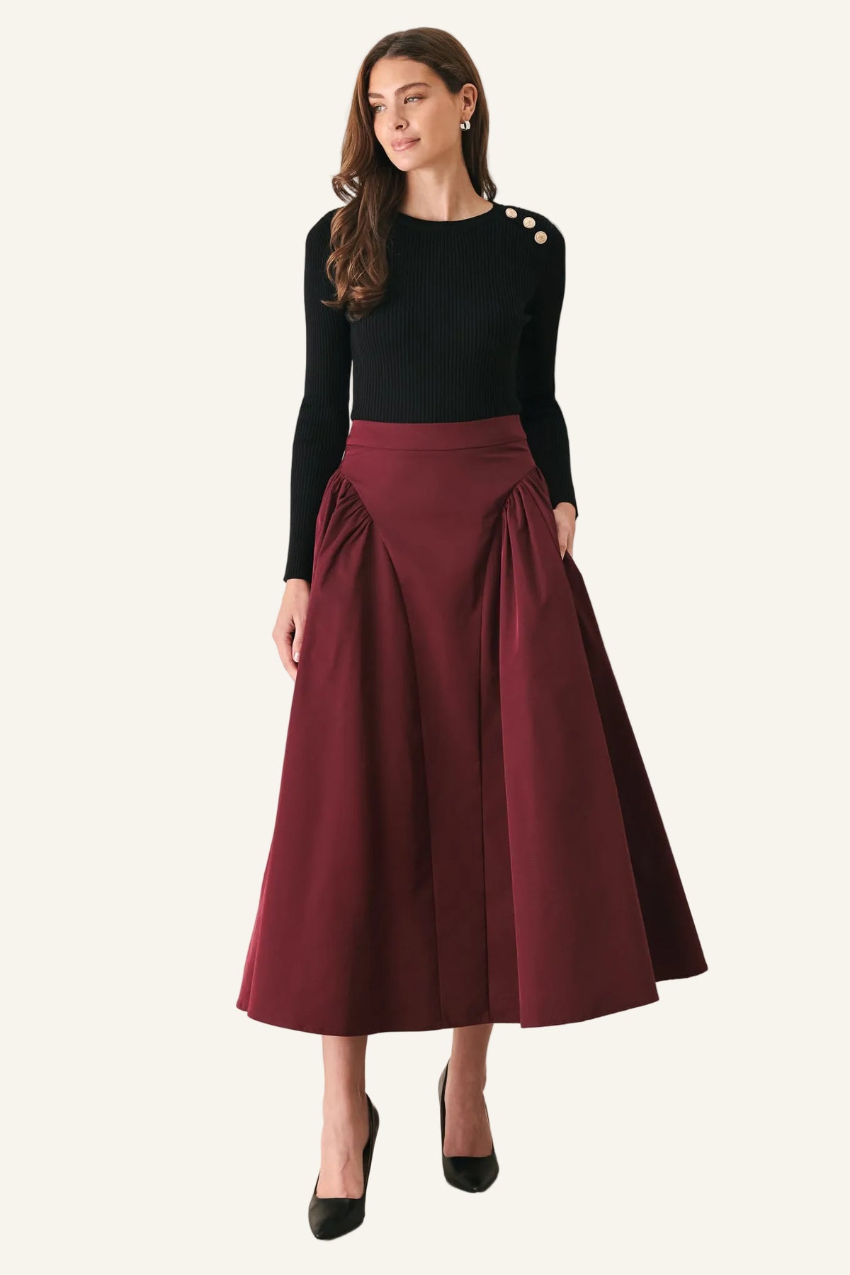 A woman modeling a black long-sleeve top with decorative gold buttons on the shoulder and a high-waisted, pleated burgundy midi skirt, paired with black heels, against a plain light gray studio background.