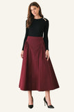 A woman modeling a black long-sleeve top with decorative gold buttons on the shoulder and a high-waisted, pleated burgundy midi skirt, paired with black high heels, against a plain light gray studio background.