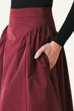 A close-up of a high-waisted, voluminous skirt in deep burgundy with a gathered waistband and side pocket, worn by a model with a black ribbed long-sleeve top.