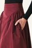 A close-up of a high-waisted, voluminous skirt in deep burgundy with a gathered waistband and side pocket, worn by a model with a black ribbed long-sleeve top.