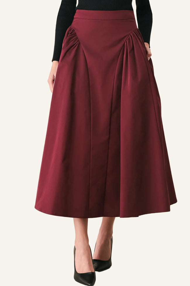 A woman wearing a high-waisted, pleated midi skirt in deep burgundy, paired with a black long-sleeve top and black pointed-toe heels, standing against a light gray studio background.