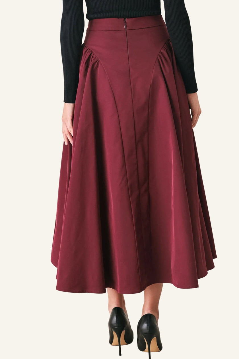 A model from the waist down wearing a high-waisted, pleated A-line midi skirt in burgundy, paired with a black long-sleeve top and black high heels.