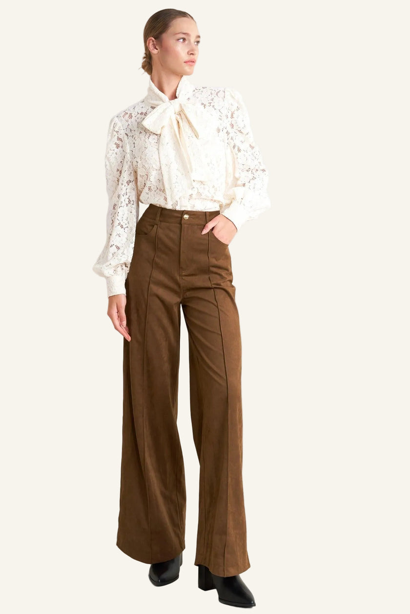 Woman wearing a white blouse with lace details and brown high-waisted pants on a white background