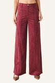A model is wearing a pair of high-waisted, wide-leg trousers in a deep burgundy corduroy fabric, paired with brown pointed-toe flats, against a plain light gray studio background.