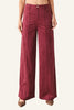 A model is wearing a pair of high-waisted, wide-leg trousers in a deep burgundy corduroy fabric, paired with brown pointed-toe flats, against a plain light gray studio background.