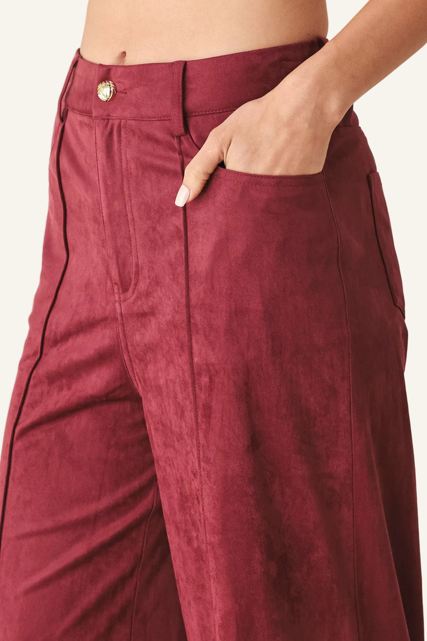 A close-up of a person wearing burgundy wide-leg trousers with black piping along the seams and a gold button at the waistband, with one hand in the pocket.