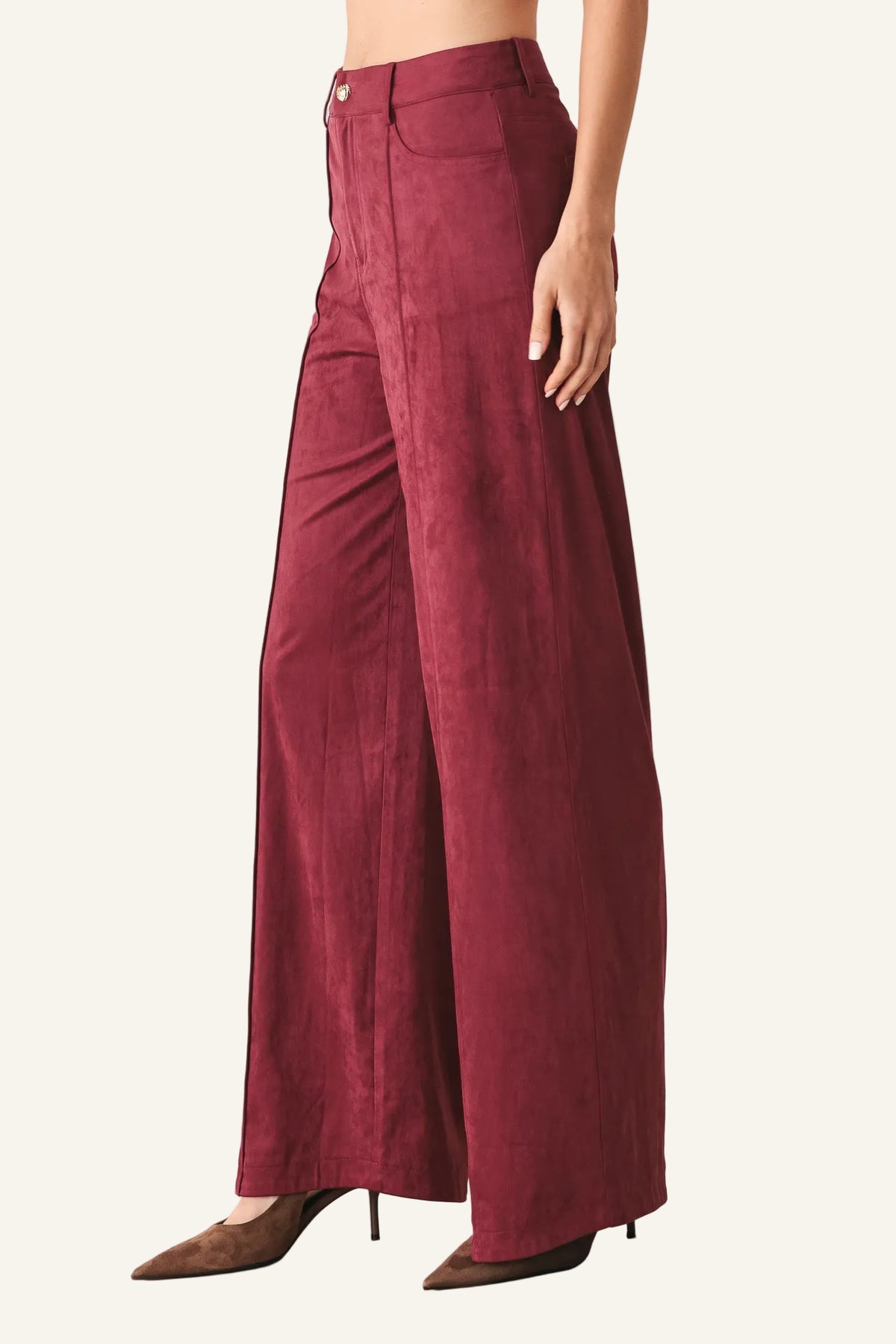 A model is wearing a pair of high-waisted, wide-leg trousers in a deep burgundy or maroon corduroy-like fabric, paired with brown pointed-toe heels, against a plain light gray studio background.