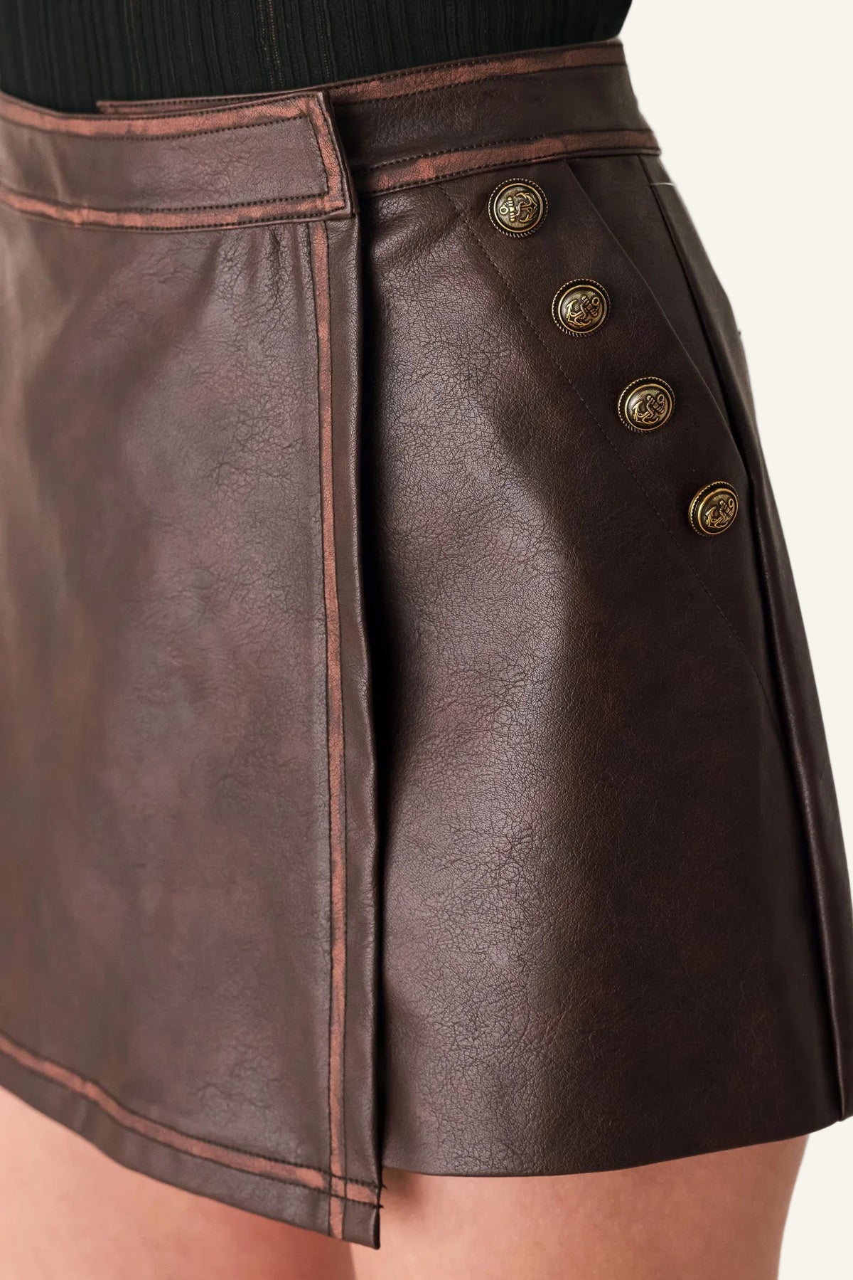 Close-up of a dark brown faux-leather wrap mini skirt with decorative gold-tone buttons on the side, shown on a model.